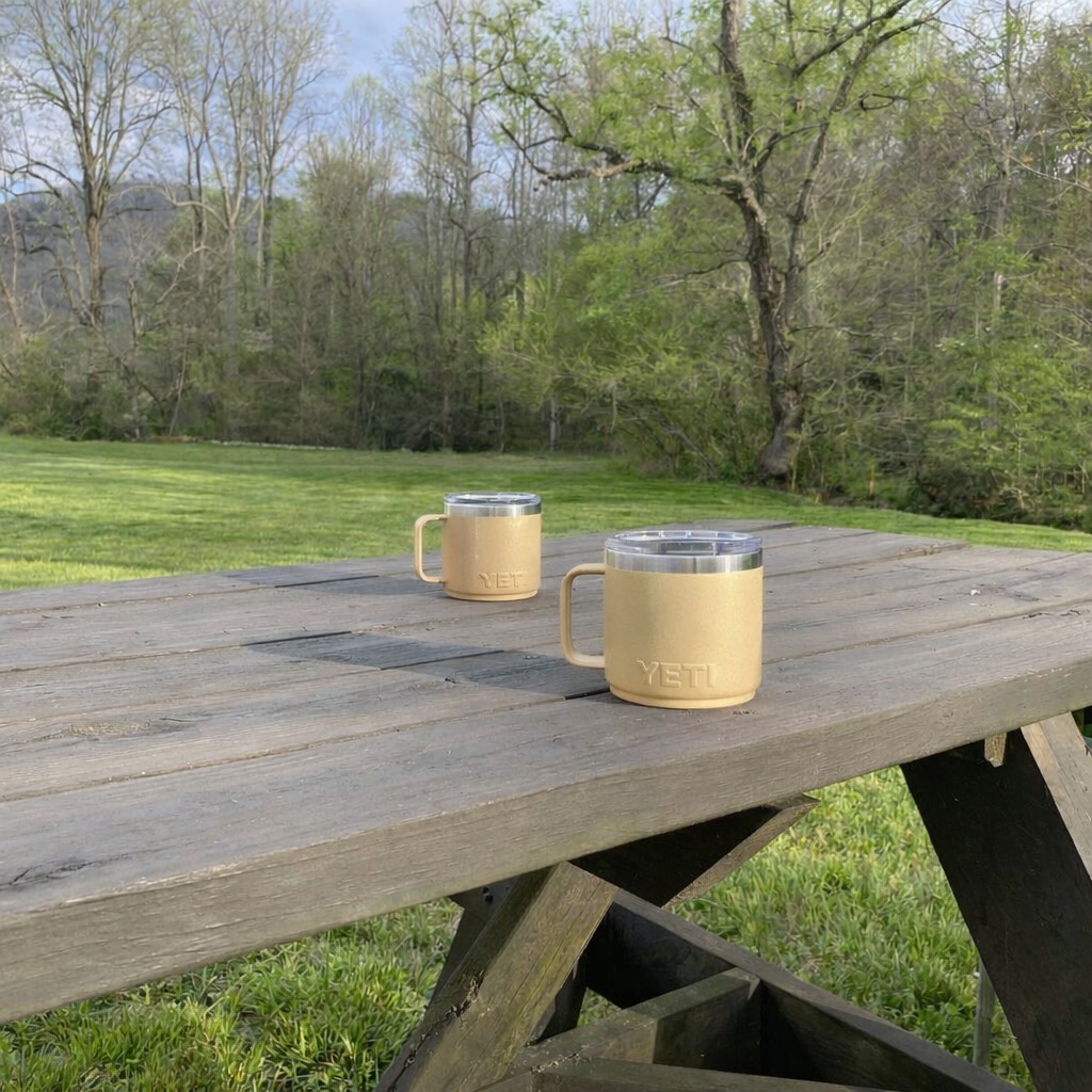 Two coffee mugs on a weathered picnic table overlooking a green meadow and spring trees in Western North Carolina